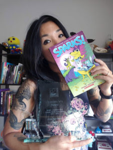 A photo of Nina Matsumoto holding up a glass trophy shaped like the province of British Columbia with her right arm, a copy of Sparks Double Dog Dare in her left hand.