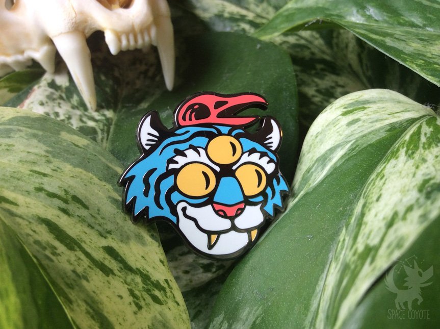 Enamel pin of a blue tiger with 3 eyes, wearing a red raven skull on its head.