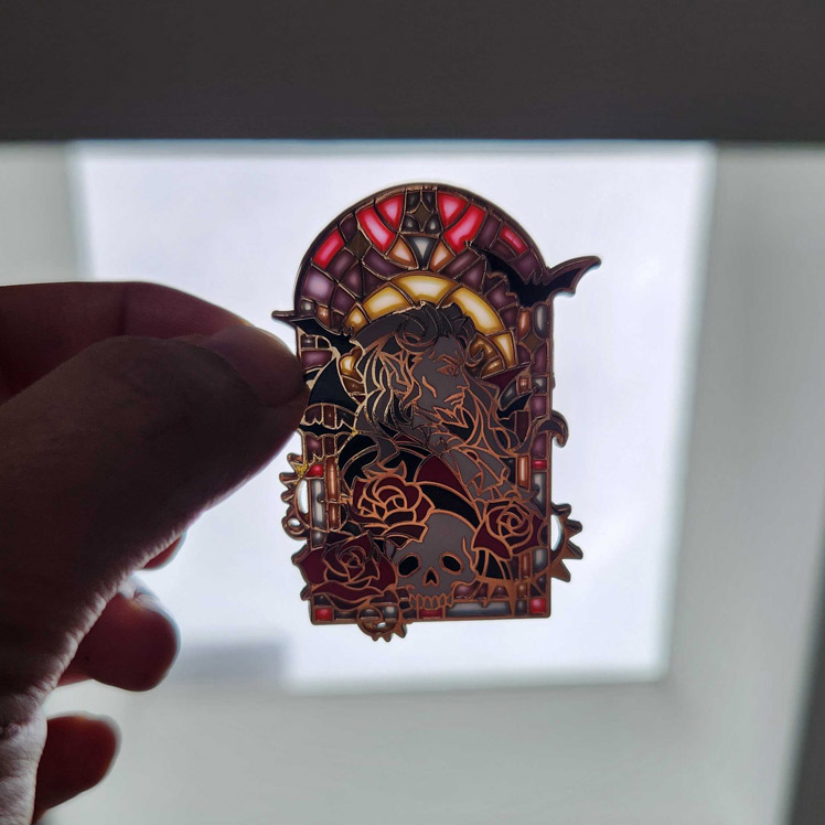 Photo of the Dracula enamel pin being held up to a light, showing the transparent stained glass effect.